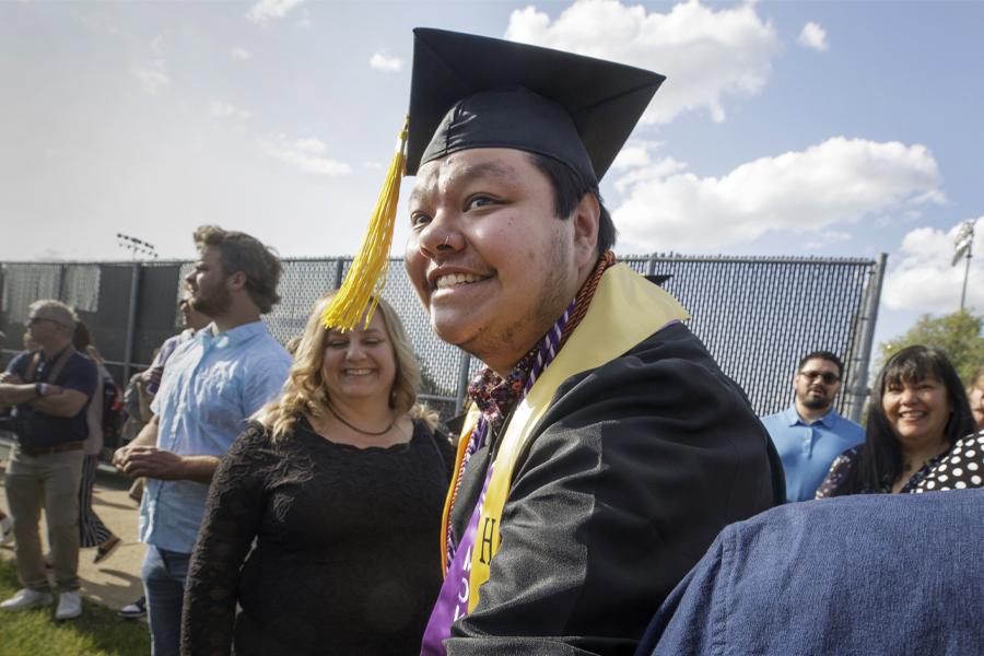 A student wearing graduate cap and gown smiles outdoors.